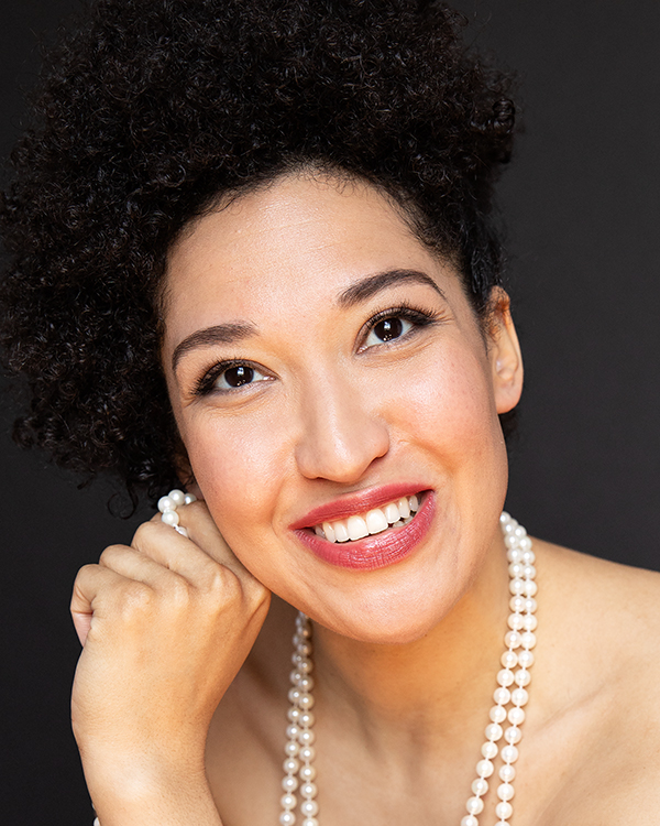 Julia Bullock, a woman with light olive skin and dark curly hair tied up, wearing a pearl necklace, smiles into the camera.