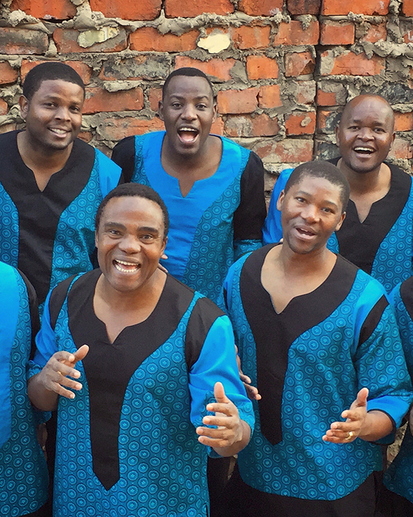 The Ladysmith Black Mambazo group of South African singers stand against a brick wall with their hands extended towards the camera, in matching colorful blue attire.