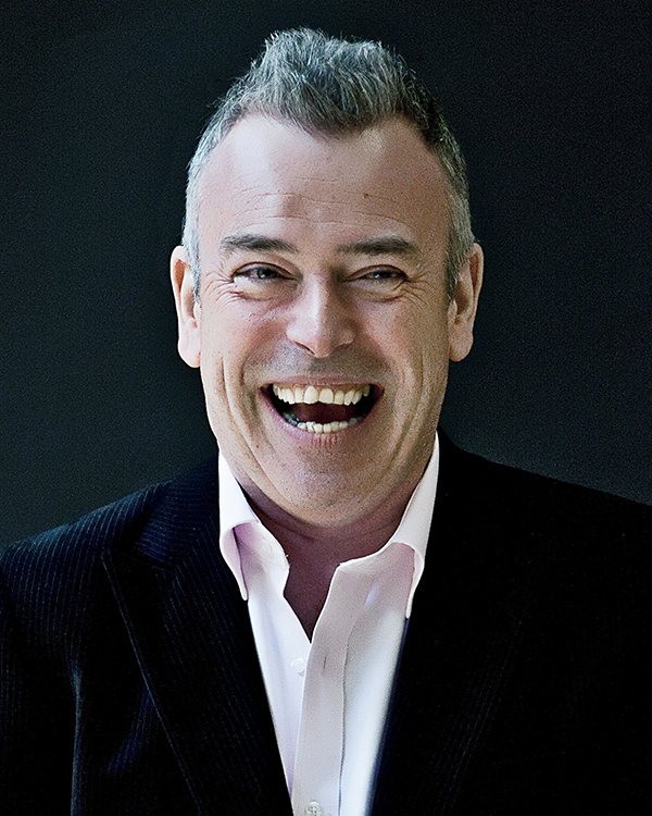 Malcolm Martineau, a white man with short salt and pepper hair, wearing a dark suit smiling widely while looking into the camera.