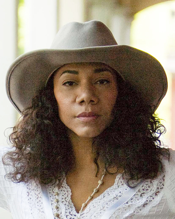 Martha Redbone, a medium-dark-skinned woman with dark curly hair looks straight into the camera wearing a beige wide-brim hat