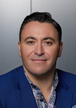 Maxim Vengerov, a white man with short dark hair slicked to one side wearing a blue suit and floral shirt, smiles into the camera