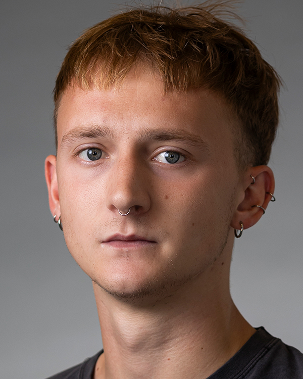 Nathan Chipps, a light-skinned man with short red hair, multiple ear piercings and a septum piercing, looks into the camera