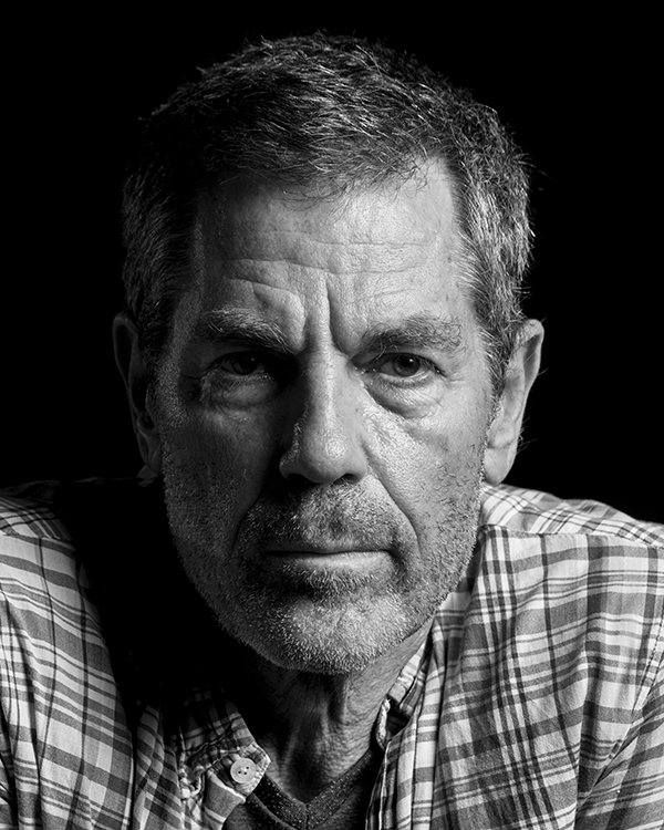 A black-and-white headshot of Ohad Naharin, an older man with salt-and-pepper hair wearing a plaid shirt, staring into the camera