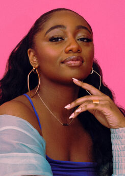 Samara Joy, a young black woman with long wavy hair wears a bright blue dress and overshirt that contrasts vividly with the hot pink background.