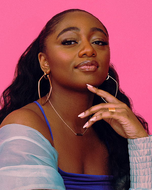 Samara Joy, a young black woman with long wavy hair wears a bright blue dress and overshirt that contrasts vividly with the hot pink background.