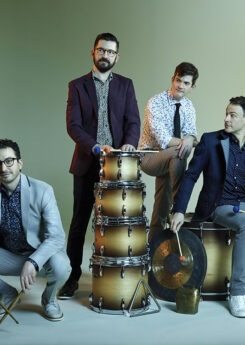 The four members of Third Coast Percussion posing with various drums and percussion instruments, two sitting and the other two standing.