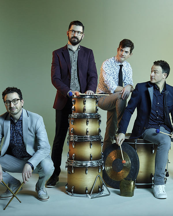 The four members of Third Coast Percussion posing with various drums and percussion instruments, two sitting and the other two standing.