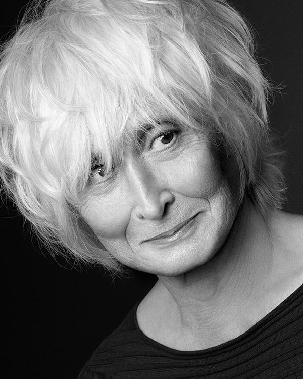 A black-and-white, close up image of Twyla Tharp smiling softly, with short white hair in a black long sleeve top.