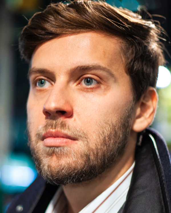 Vladimir Rumyantsev, a Russian man with short brown hair wearing a striped button-up and black coat, looks off to the side of the camera.