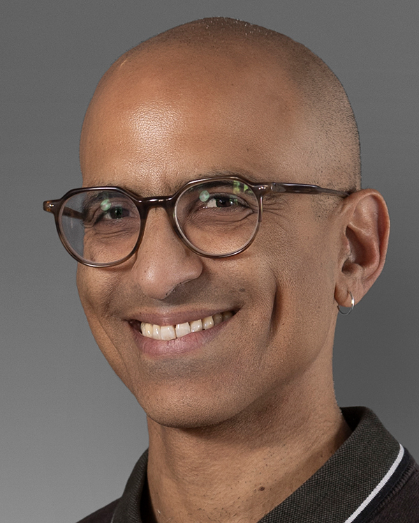 Yaniv Nagar, a tan man with a shaved head wearing round brown glasses, smiles widely at the camera