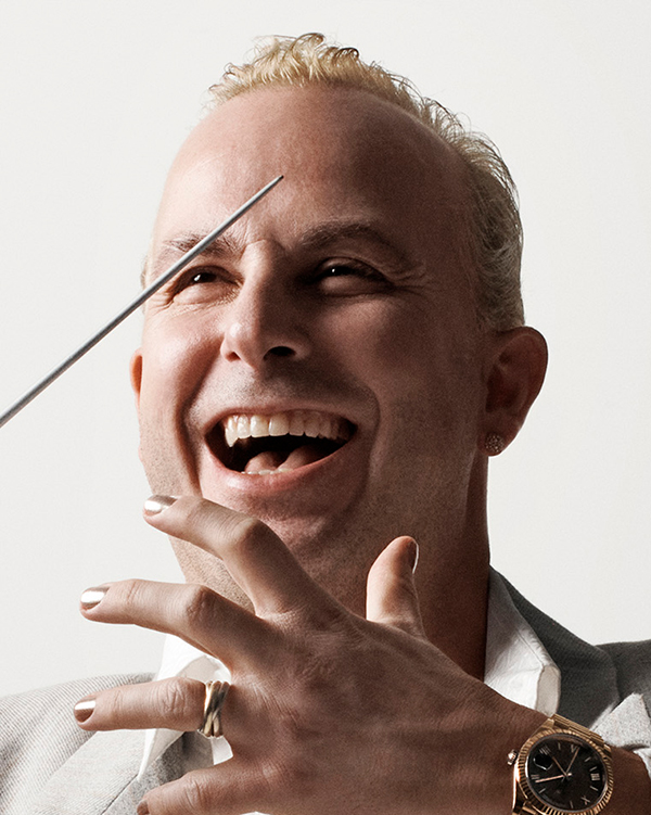 Yannick Nézet-Séguin, a white man with short blond hair in a grey suit, smiles while posing with his conductor's baton.