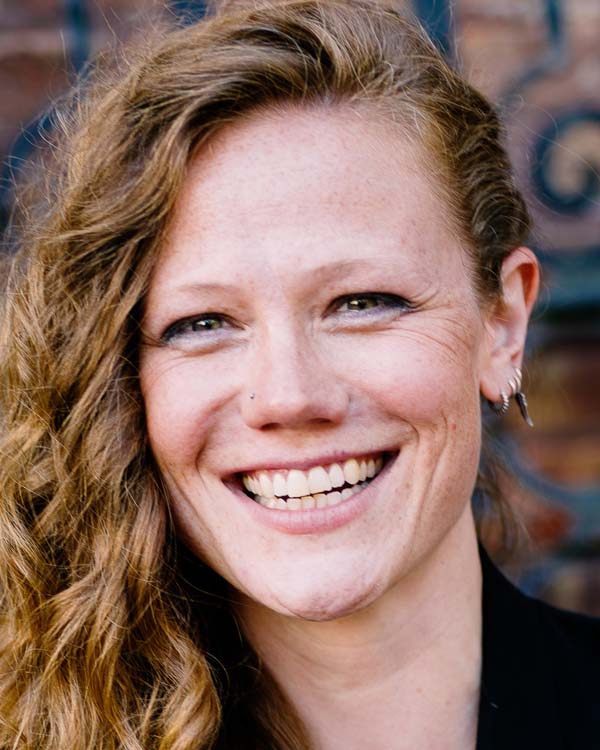 Constance Volk, a white woman with long curly brown hair, smiles widely looking into the camera.