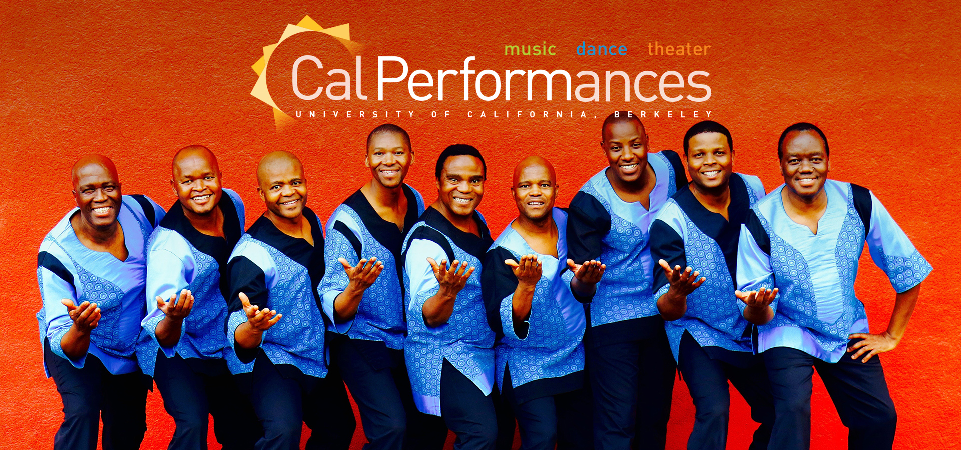 The Ladysmith Black Mambazo group of South African singers stand against a bright red wall with their hands extended towards the camera, in matching colorful blue attire.