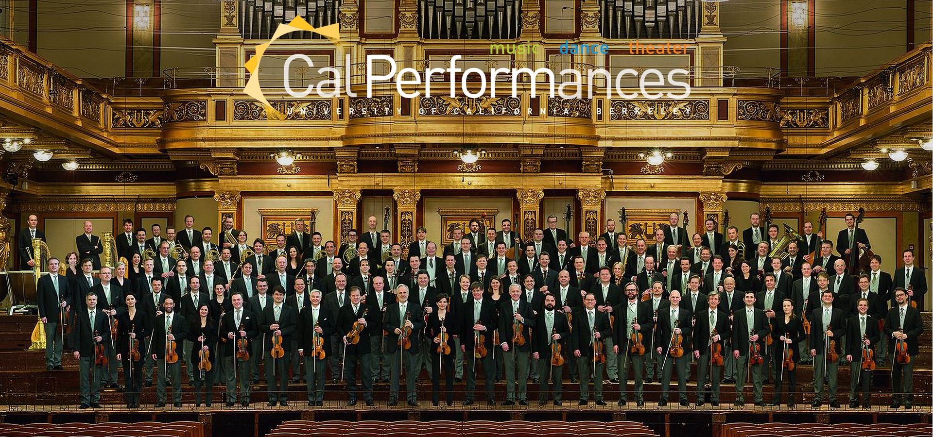 The Vienna Philharmonic stand as a large group in front of an ornate gold organ, holding their instruments and wearing formal dress.