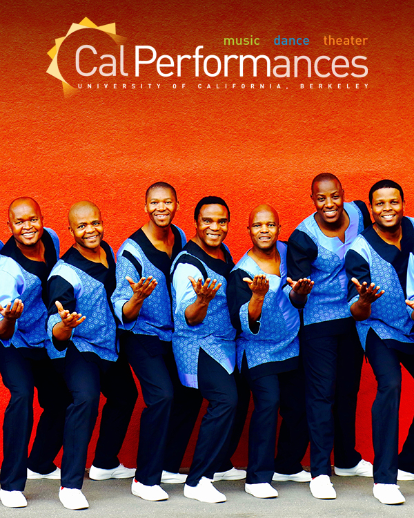 The Ladysmith Black Mambazo group of South African singers stand against a bright red wall with their hands extended towards the camera, in matching colorful blue attire.