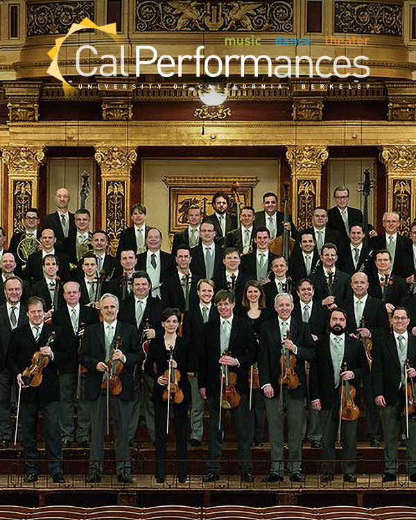 The Vienna Philharmonic stand as a large group in front of an ornate gold organ, holding their instruments and wearing formal dress.