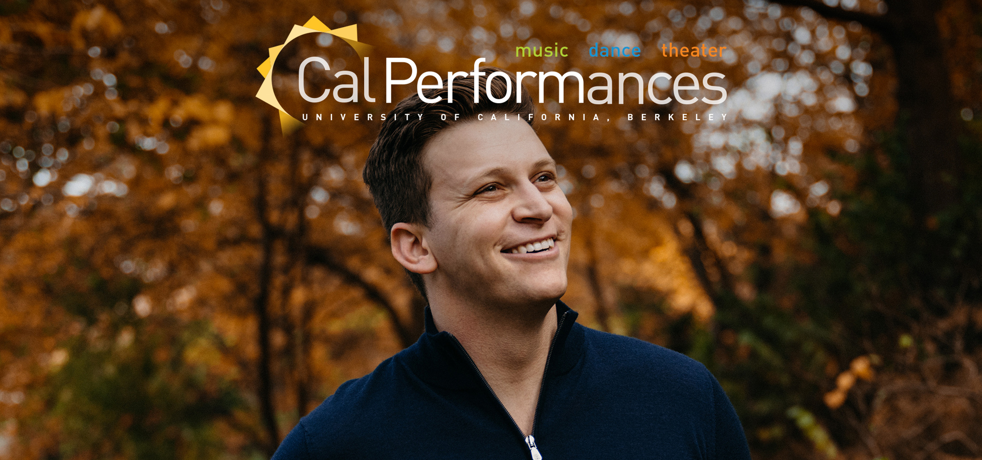 Aryeh Nussbaum, a young white man with short brown hair, wears a blue sweater smiling to his left in front a background of trees with orange leaves.