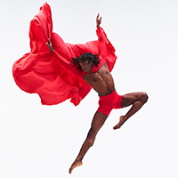 Dancer from the the Alvin Ailey American Dance Theater poses against a white background in a powerful jump wearing vibrant red shorts and a red cape.