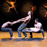 Four bare-chested male dancers in the Batsheva Dance group balance upon one another in an on-stage performance of MOMO.