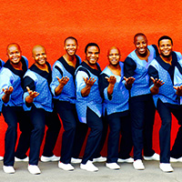 The Ladysmith Black Mambazo group of South African singers stand against a bright red wall with their hands extended towards the camera, in matching colorful blue attire.