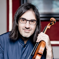 Leonidas Kavakos, a white man with dark shoulder-length hair and glasses, holds his violin and looks into the camera.