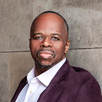 Lester Lynch, an African American opera singer, poses against a stone background wearing a plum jacket and white button-down shirt.