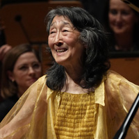 Pianist Mitsuko Uchida stands smiling in sparkling yellow attire, in front of her piano with the Mahler Chamber Orchestra behind her.