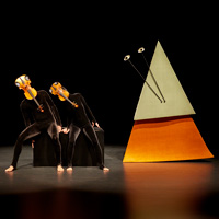 Performers in all-black outfits dance with decorated violins over their faces, next to a bright sculpture on stage.