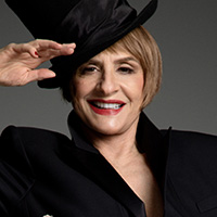 Patti LuPone smiles at the camera wearing statement red lipstick, outfitted in a bold black suit and shiny black top hat.