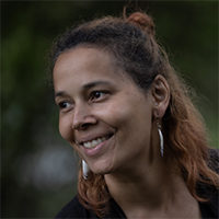 Rhiannon Giddens, a woman with long reddish-dark hair, sits looking to the left of the camera in a black top and holding a banjo.