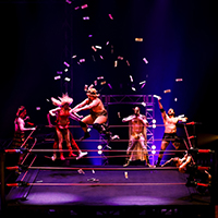 Two Cirque FLIP Fabrique performers are dressed as pro-wrestlers in the center of a wrestling ring, and meet airborne in a forceful acrobatic collision with each other.