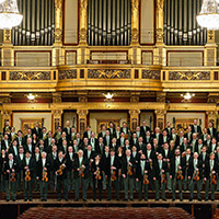 The Vienna Philharmonic stand as a large group in front of an ornate gold organ, holding their instruments and wearing formal dress.