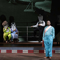 Performers in elaborately patterned clothing stand facing the audience with colorful projected words and shapes in the background.