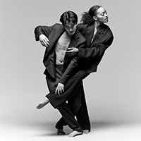 A black-and-white image of two dancers of the A.I.M. company wear slacks, blazers, and opened button-ups making an intricate pose with their arms.