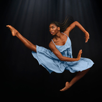 A black female dancer of the Alvin Ailey American Dance Theater with long braided hair and wearing a light blue dress in a jump pose against a dark background