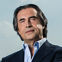  Riccardo Muti stands at a podium with his baton in hand, conducting with a serious expression on his face.