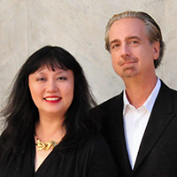 Wu Han, an Asian woman long dark hair, and David Finckel, a white man with gray-brown hair, stand side-by-side wearing formal attire as they smile into the camera.