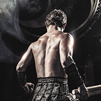A male drummer of Drum Tao stands with his back facing the camera as he stares at a large taiko drum with a foggy gray background.