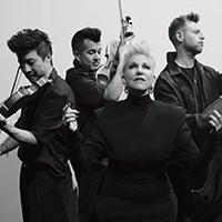 A black-and-white image of Joyce DiDonato, Kevin Puts, and the members of Time for Three stand around a large box that reads, "Emily No Prisoner Be."