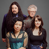 The four members of the Kronos Quartet, two men and two women, wear small smiles as they sit in front of a gray background.