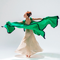 A dancer from Martha Graham Dance Company in a white long dress and flowing green cape trimmed with black posed against a very light background