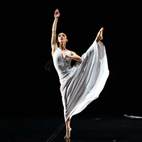 A dancer of Martha Graham Dance Company in a white flowy dress posed with her left leg and right arm up in the air.
