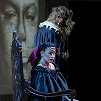 The lead actors of Opera Parallele's La Belle et La Bête perform onstage, with La Bête in the background, gazing at Belle as she sits in a chair, wearing a somber expression.