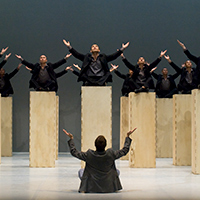The monks of the Shaolin Temple sit atop tall wooden blocks onstage, looking upwards with their arms outstretched.