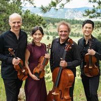 The Takács Quartet members, three men and one woman, stand smiling with their instruments in hand in front of a lush forest background.