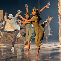 Dancers of the Joffrey Ballet wear flowy earth toned outfits onstage in matching poses, holding prop wineglasses in hand.