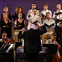 Conductor Masaaki Suzuki leads the Monteverdi Choir, a group of men and women, as they perform for an audience in a concert setting.