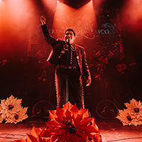A man dressed in a mariachi outfit performs on stage, adorned with flowers and bathed in red light, celebrating the culture of Mariachi Herencia de México.